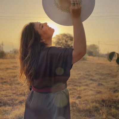 Mulher segurando chapéu bege decorado com flores em campo ao pôr do sol