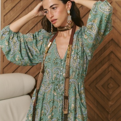 Vestido comprido verde com padrão paisley usado por mulher com acessórios em ambiente interior com parede de madeira e sofá bege.