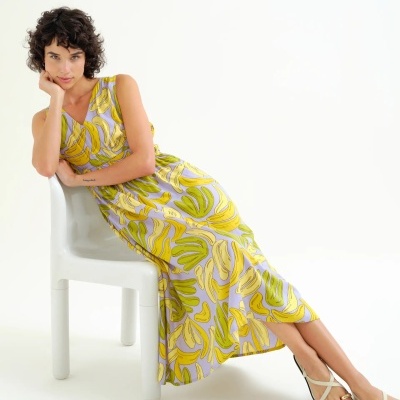 Vestido comprido com padrão de folhas amarelas e verdes, mulher sentada em cadeira branca com sandálias brancas.