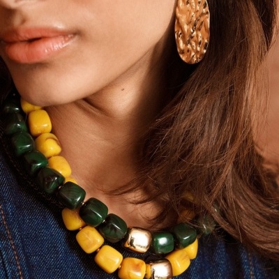 Colar de contas amarelas, verdes e douradas com brinco dourado texturizado num pescoço feminino