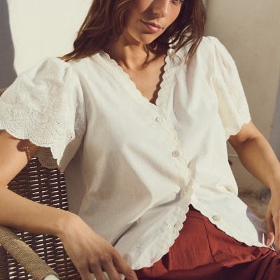 Blusa branca com botões e mangas bordadas, mulher sentada em cadeira de vime.