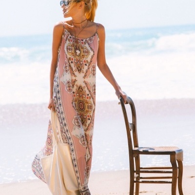 Mulher com vestido comprido e padrão floral segurando saco branco ao lado de cadeira de madeira na praia.