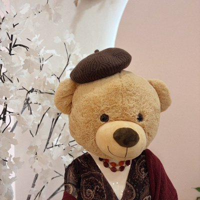 Ursinho de peluche com chapéu castanho, vestido com blusa castanha e xale vinho sentado em cadeira rosa.