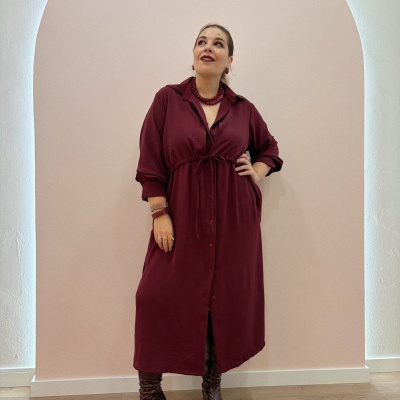 Mulher a usar vestido comprido vinho com cinto e botões, fundo parede bege com arco e chão de madeira