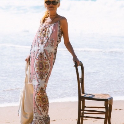 Vestido comprido com padrão floral e geométrico em tons suaves com alças finas junto a cadeira de madeira na praia