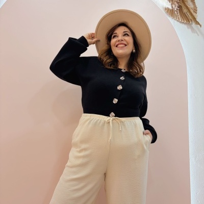 Mulher com camisa preta e calças creme a usar chapéu bege em fundo rosa claro com arco e flores secas.