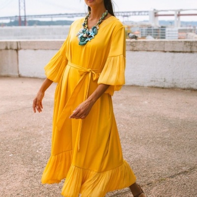 Mulher com vestido amarelo comprido e colar grande colorido em ambiente exterior com uma ponte ao fundo.