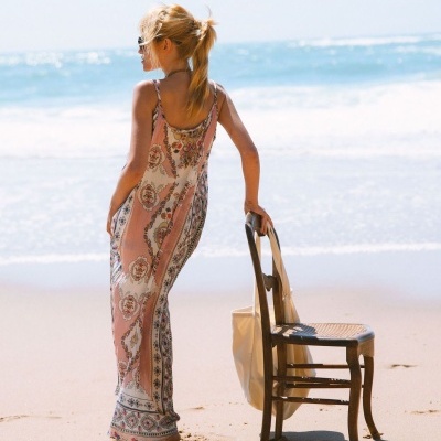 Mulher na praia com vestido longo rosa e branco segurando uma cadeira de madeira com bolsa de tecido.