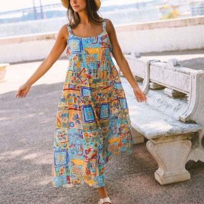 Mulher com vestido colorido comprido, chapéu de palha e sandálias brancas ao ar livre junto a banco de pedra.