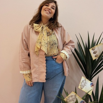 Mulher com blusa amarela floral, casaco rosa e calças de ganga azuis junto a plantas com cartões