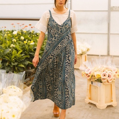 Mulher com vestido comprido azul com padrões geométricos a usar t-shirt branca e sandálias castanhas, num local com flores e caixas de plástico.