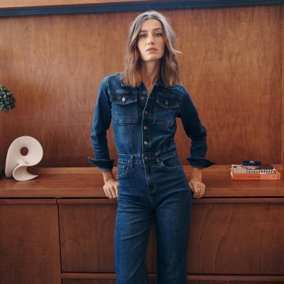 Mulher veste macacão de ganga azul escuro com botões e bolsos no peito em ambiente com móvel de madeira.