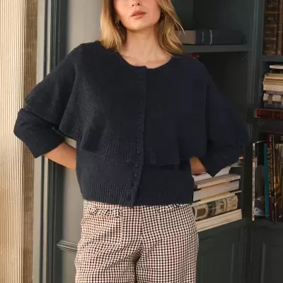 Mulher vestindo cardigan azul escuro de malha com botões e calças xadrez num ambiente com estantes de livros.