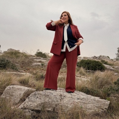 Mulher com fato feminino vermelho, camisa branca com riscas, gravata branca e colete azul escuro em campo rochoso