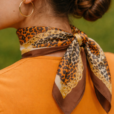 Pessoa com lenço animal print castanho e amarelo no pescoço, t-shirt laranja e brinco de argola dourado