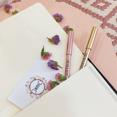 Duas canetas metalizadas, flores secas coloridas, folha branca com selo IMPRENSA, tapete rosa com padrão branco