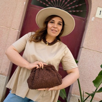 Mulher com blusa bege, chapéu e bolsa de crochet castanha em frente a porta vermelha com arco.