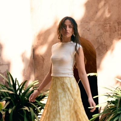 Mulher com camisola branca e saia amarela num pátio com plantas.