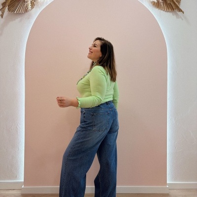 Mulher com camisola verde e calças de ganga azuis largas em fundo rosa com arco e decoração floral seca