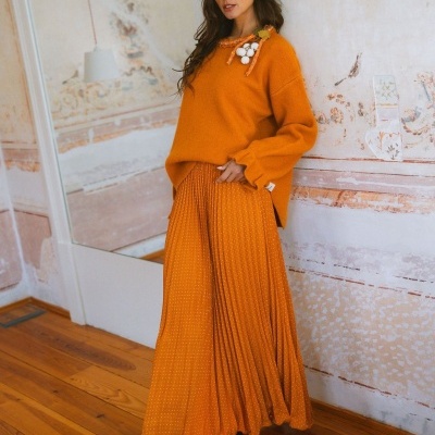 Mulher com suéter laranja e saia plissada laranja em interior com chão de madeira.