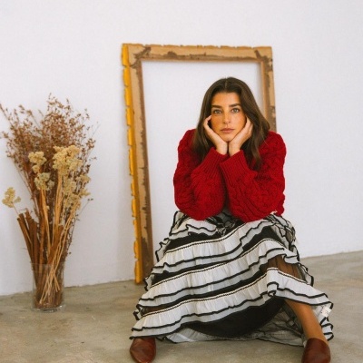 Mulher com camisola vermelha e saia branca com riscas pretas sentada em chão de cimento com quadro de madeira e vaso de flores secas ao fundo