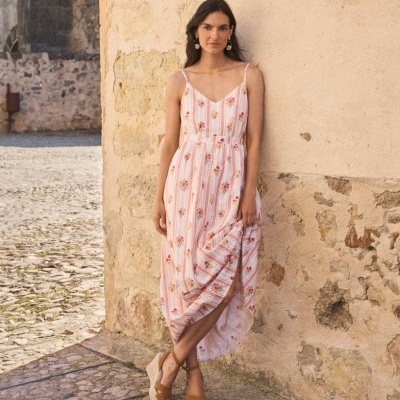 Mulher com vestido comprido listrado rosa com flores encostada numa parede de pedra