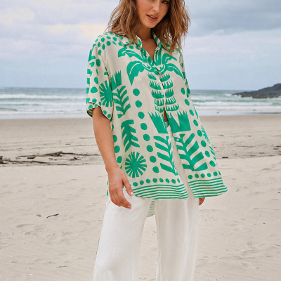 Mulher de blusa verde e branca com padrão tropical e calças brancas na praia