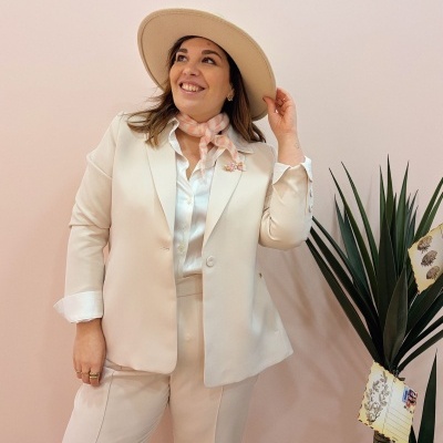 Mulher com fato de senhora creme, camisa branca, lenço rosa, chapéu creme e broche, junto a planta verde com cartões