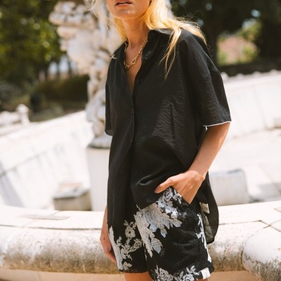 Mulher com óculos redondos castanhos e roupa preta com padrão floral branco ao ar livre