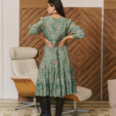 Mulher de costas com vestido verde floral e botas pretas em ambiente interior com cadeira e painéis de madeira.