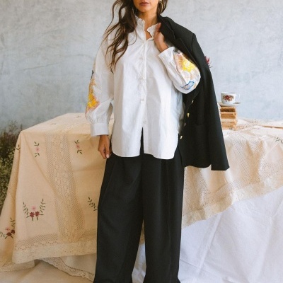 Mulher com camisa branca bordada, calças pretas largas e blazer preto sobre ombro em ambiente com toalha bordada.