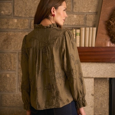 Blusa verde-oliva com bordado floral, vista de costas, contra parede de pedra e prateleira com livros