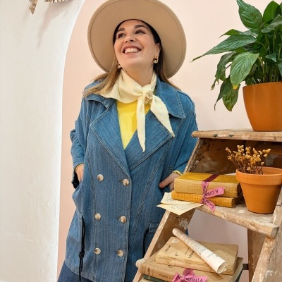 Mulher com casaco azul denim, lenço amarelo e chapéu bege ao lado de escada de madeira com livros e plantas