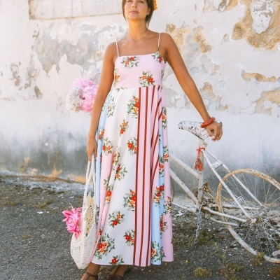 Mulher a usar vestido comprido com padrão floreado e riscas, bolsa de crochet e sapatos castanhos, com bicicleta antiga e parede rústica ao fundo.