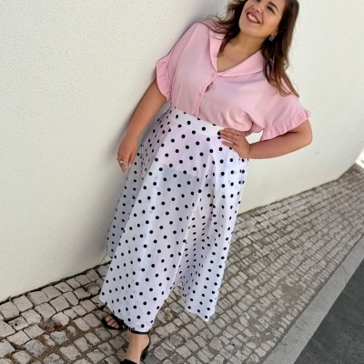 Pessoa com blusa rosa e saia branca com bolinhas pretas junto a parede branca.