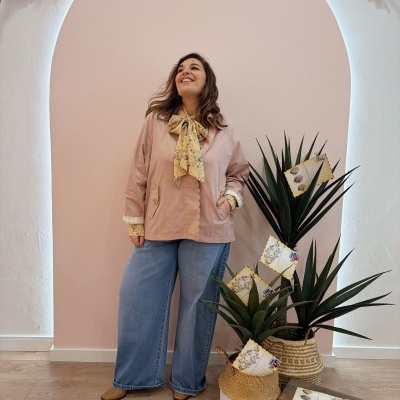 Mulher com blusa rosa com laço amarelo e calças de ganga, parede rosa com arco e plantas em cestos de palha