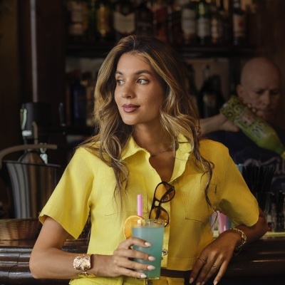 Mulher de camisa amarela e calças altas segurando bebida azul num bar