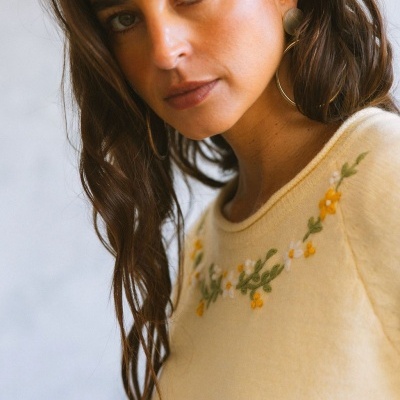 Suéter amarelo claro bordado com flores, mulher com brincos dourados e cabelo castanho