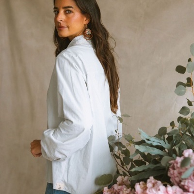 Camisa branca de tecido leve com etiqueta maitté visível na barra, usada por mulher com cabelo castanho e calças de ganga azuis, cenário com flores rosas e folhas verdes.