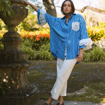 Mulher com camisa azul às riscas brancas, calças brancas e sapatos castanhos em jardim.