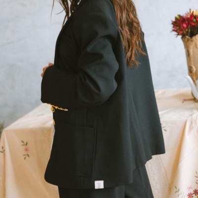 Blazer preto com botões dourados e bolsos, em ambiente interior com mesa e flores