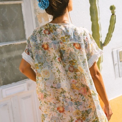 Mulher a usar túnica floral leve e transparente com renda nas mangas, cabelo apanhado com flor azul, parede branca e cacto ao fundo