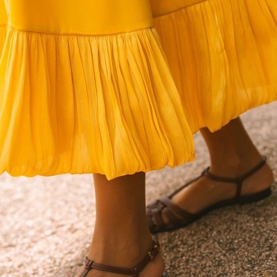 Vestido amarelo com bainha plissada e sandálias castanhas em chão de cascalho