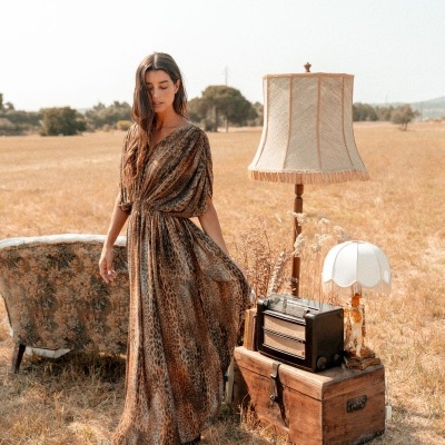 Mulher com vestido longo animal print num campo seco junto a móveis vintage e luminárias.