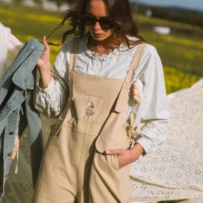 Macacão bege com bordado floral e renda sobre blusa branca num campo com flores amarelas