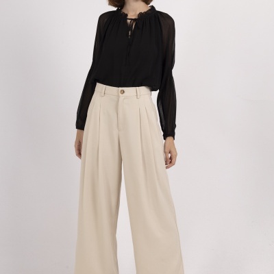 Mulher com blusa preta transparente e calças largas creme num fundo branco.