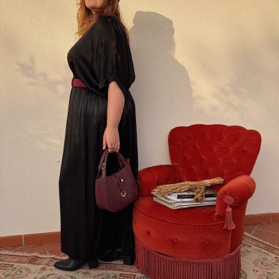 Mulher com vestido preto e chapéu preto com adereço dourado, bolsa castanho-avermelhada ao lado de cadeira vermelha.