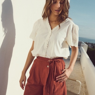 Mulher com blusa branca de mangas bufantes e calções terracota de linho na varanda com vista para o mar