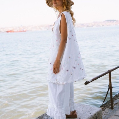 Mulher com vestido branco bordado, chapéu de palha e sandálias junto ao mar
