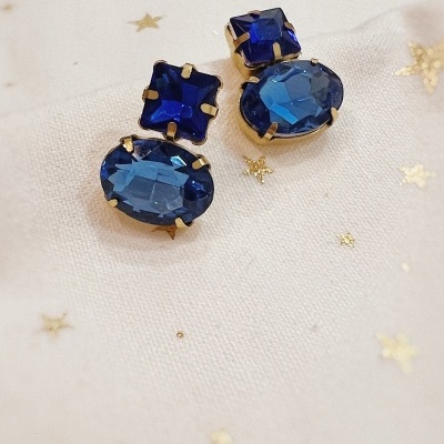 Brincos com pedras azuis facetadas em metal dourado sobre tecido branco com estrelas douradas.